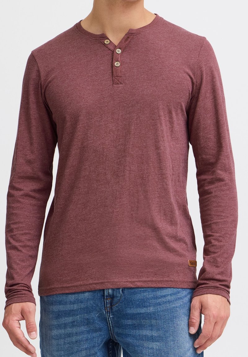 Homme portant un t-shirt henley bordeaux à manches longues avec trois boutons et un jean bleu, debout devant un fond uni.