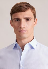 Jasnoniebieska koszula z delikatnym geometrycznym wzorem, zapinana na guziki z klasycznym kołnierzykiem. Gładka tekstura.