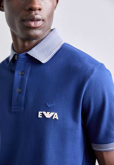 Zils polo krekls, kas izgatavots no teksturētas auduma, ar baltu un tumši zilu svītru apkakli, trīs pogām un izšūtu logotipu uz krūtīm.