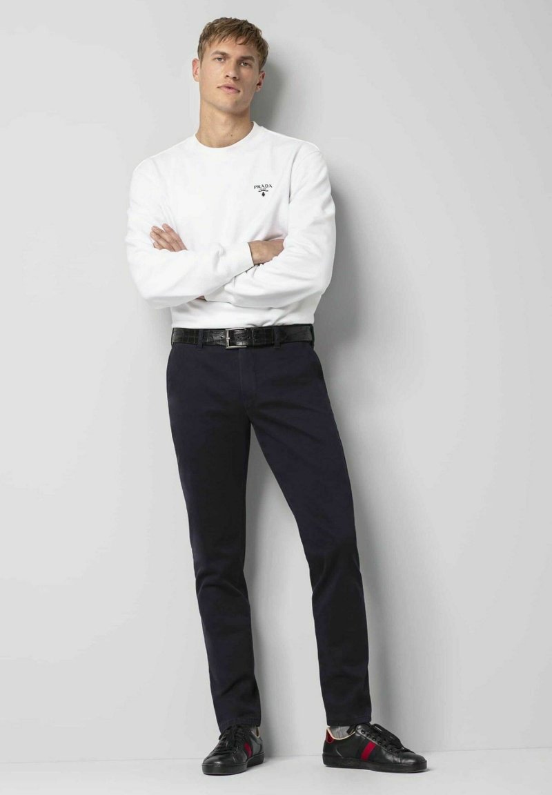 Weißer Sweatshirt mit Logo, dunkelblaue Hose und schwarze Sneakers mit roten Akzenten. Das Modell posiert mit verschränkten Armen vor einem hellen Hintergrund.