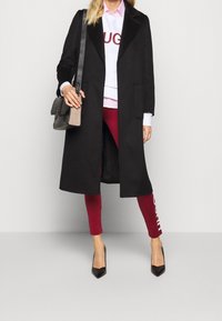 Svart ullkappa med slagkrage, bärs över en vit sweatshirt och en rosa skjorta. Röda leggings med en vit logotyp. Svarta spetsiga klackar och en grå väska.