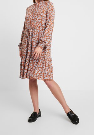 Langwerpige jurk in roestkleur met een bloemenpatroon in blauw en wit. Bevat een hoge hals, elastische manchetten en een uitlopende rokontwerp. Gestyled met zwarte loafers.