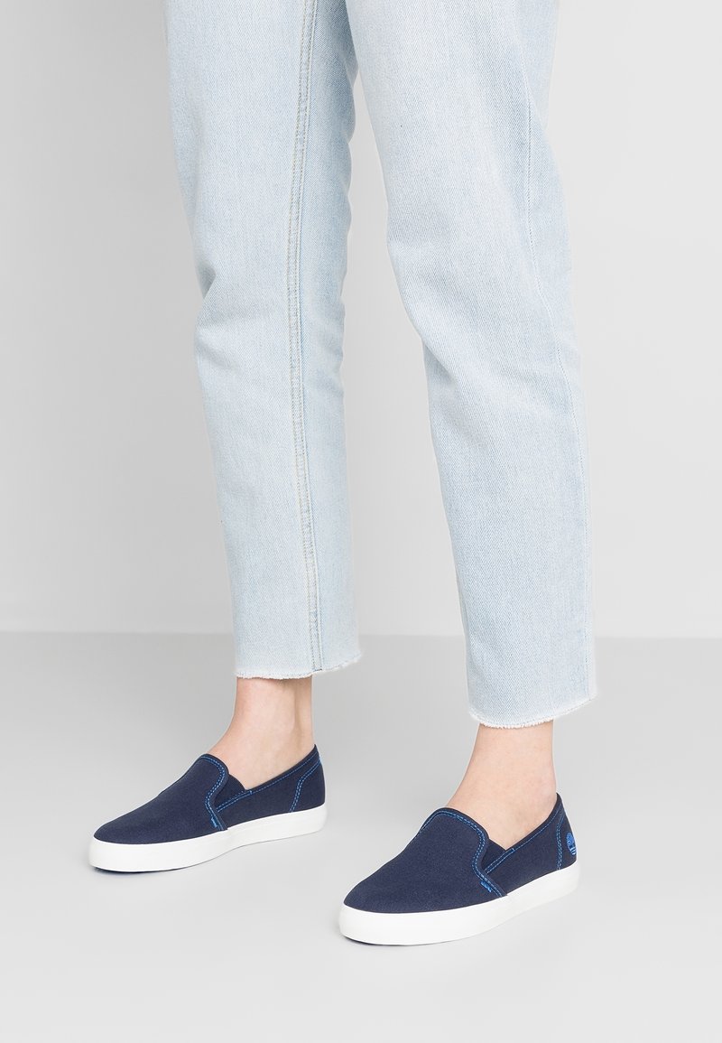 Chaussures en toile bleu marine à enfiler avec semelles en caoutchouc blanc et détails de couture bleue, assorties à un jean court bleu clair.
