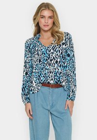 Långärmad blus i blå och svart leopardmönster på en vit bakgrund, med krage och lös passform, kombinerad med högmidjade jeans.