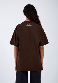 Camiseta de algodón marrón con un ajuste relajado, mangas cortas y cuello redondo. Cuenta con un logo de firma blanco en la parte superior de la espalda.