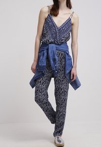 Navy patterned jumpsuit with spaghetti straps and a v-neck, paired with a denim shirt tied around the waist and metallic shoes.