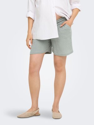 Person iført lysegrønne teksturerede shorts, hvid skjorte med knapper og beige vævede slingback-flats med den ene hånd i lommeposen på shortsene.