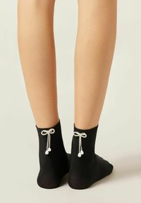 Sorte ankelstrømper med en glat tekstur, der har dekorative sløjfer bagpå med hængende krystal- og perleudsmykninger.