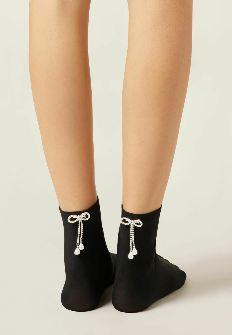 Sorte ankelstrømper med en glat tekstur, der har dekorative sløjfer bagpå med hængende krystal- og perleudsmykninger.