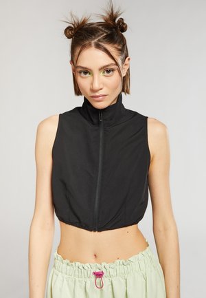 Gilet nero cropped con colletto alto e zip frontale, realizzato in materiale leggero. Il modello indossa pantaloni elastici verde chiaro con dettagli rosa.