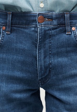 Close-up van de taille van blauwe spijkerbroeken met een koperen knoop en riemlussen over een ingestopt lichtblauw overhemd.