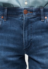 Κοντινό πλάνο στη μέση ενός μπλε τζιν με χαλκό κουμπί και θηλιές ζώνης πάνω από ένα ανοιχτό μπλε πουκάμισο που έχει μπει μέσα.