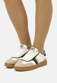 Weiße und beige Sneaker mit schwarzen Akzenten, Gummisohle, strukturiertem Design, Schnürsenkeln und Branding an der Seite. Geeignet für Freizeitkleidung.