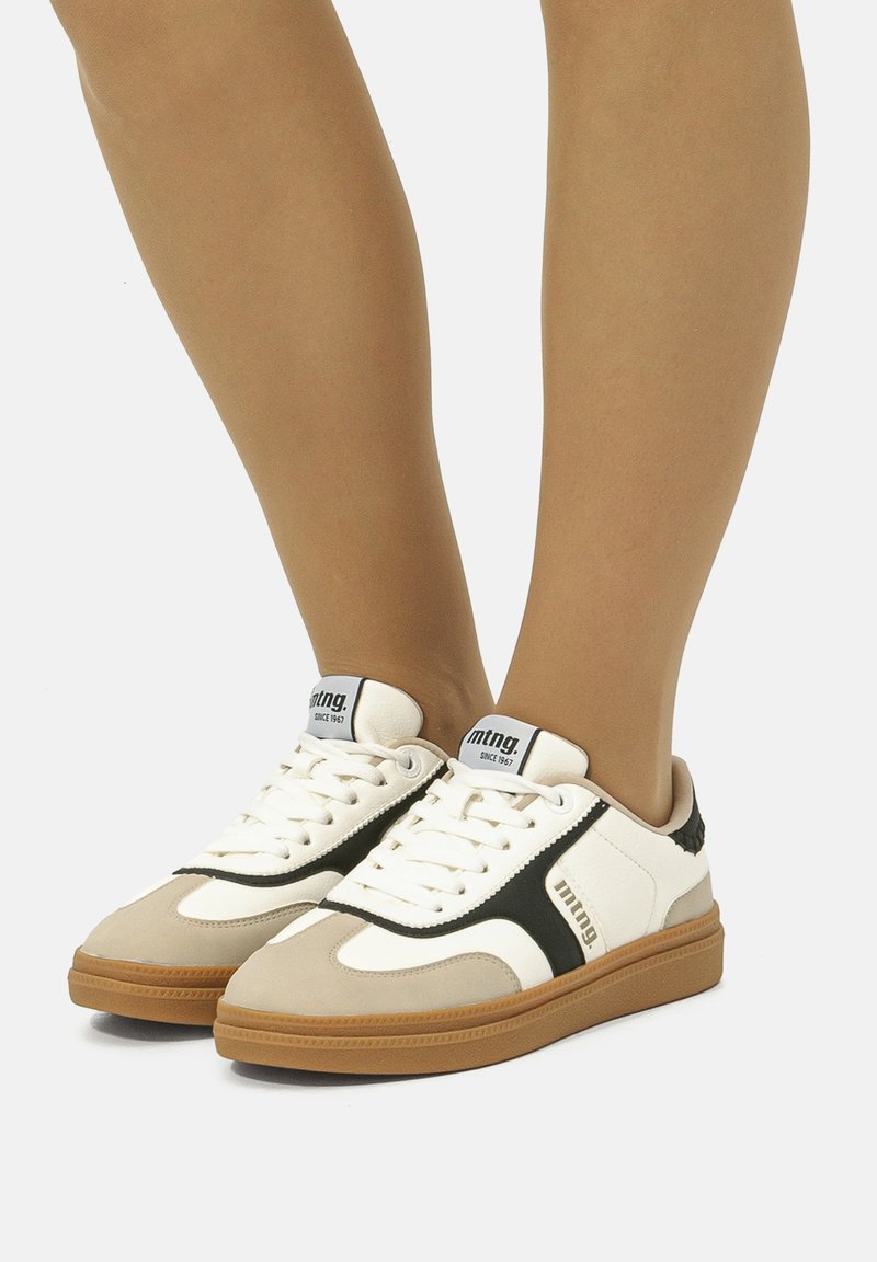 Weiße und beige Sneaker mit schwarzen Akzenten, Gummisohle, strukturiertem Design, Schnürsenkeln und Branding an der Seite. Geeignet für Freizeitkleidung.