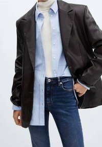 Personne portant des vêtements superposés : col roulé blanc, chemise boutonnée bleu clair, blazer en cuir marron foncé et jean bleu.