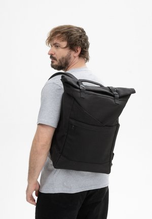 Man met baard die een lichtgrijs t-shirt en een zwarte broek draagt, draagt een grote zwarte roll-top rugzak op zijn rug en kijkt naar links.