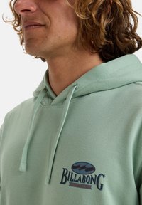 Sudadera verde menta de tejido suave, con un bolsillo frontal tipo canguro, capucha con cordón y un logo de Billabong en azul y rojo en el pecho.