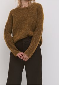 Kobieta w puszystym, brązowym sweterze i czarnych, wysokich spodniach, z dłońmi swobodnie złożonymi z przodu na jednolitym tle.