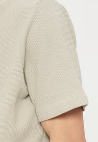 Μπεζ πλεκτό πουκάμισο με κοντά μανίκια και υφή που μοιάζει με βάφλα, εμφανίζεται στο πάνω μέρος του μπράτσου και στον ώμο ενός ατόμου.