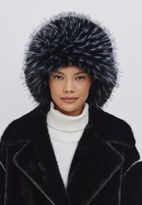Sort faux fur-hat med en fyldig, spids tekstur. Båret sammen med en hvid turtleneck sweater og en sort frakke, der fremhæver lag og kontraster.