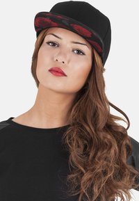 Schwarze Kappe mit floralem Muster am Schirm, aus Stoff. Getragen mit einem schwarzen Oberteil, das lange, wellige Haare zeigt.