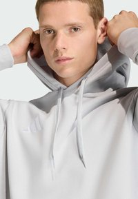 Ανοιχτό γκρι φούτερ με κουκούλα και μαλακή υφή, με μινιμαλιστικό λευκό λογότυπο και κορδόνια, δομημένη εφαρμογή και τσέπη καγκουρό.