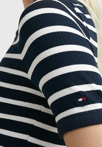 Niebiesko-białe paskowane dzianinowe bluzka o ryflowanej teksturze. Posiada krótkie rękawy i mały akcent z logo na rękawie.