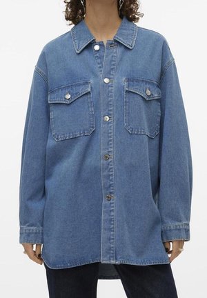 Vrouw draagt een oversized blauwe denim overhemdjas met knoopsluiting aan de voorkant en twee borstflapzakken.