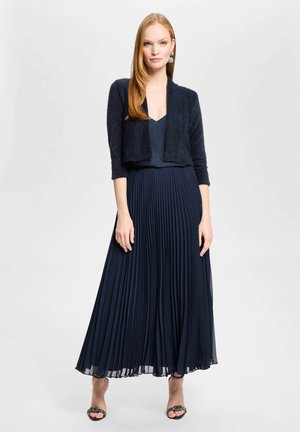 Donna che indossa un cardigan corto blu navy, gonna lunga plissettata blu navy e sandali neri con tacco su sfondo bianco.