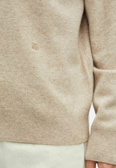 Beige gebreide trui met geribbelde zoom en boorden, voorzien van een subtiel logo. Het materiaal lijkt zacht en heeft een gestructureerde uitstraling met een licht gemêleerd effect.