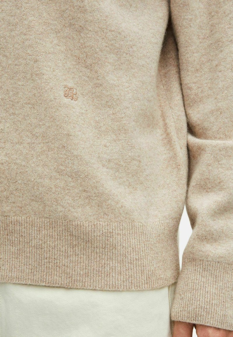 Beige gebreide trui met geribbelde zoom en boorden, voorzien van een subtiel logo. Het materiaal lijkt zacht en heeft een gestructureerde uitstraling met een licht gemêleerd effect.