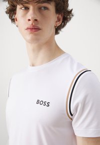 Camiseta blanca de manga corta con textura acanalada, que presenta un logotipo negro de "BOSS" y acentos de rayas marrones y negras en los hombros.