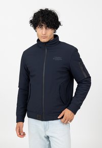 Navyblaue Zip-Up-Jacke aus weichem Material, mit hohem Kragen, zwei Fronttaschen und einer Ärmel-Tasche mit Reißverschluss.