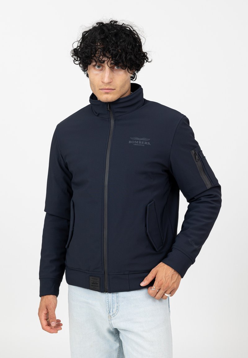 Navyblaue Zip-Up-Jacke aus weichem Material, mit hohem Kragen, zwei Fronttaschen und einer Ärmel-Tasche mit Reißverschluss.