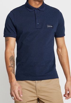 Námorně modré polo tričko vyrobené z bavlny, s límečkem, třemi knoflíky a obdélníkovou nášivkou loga Calvin Klein na hrudi.