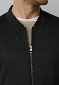 Veste bomber noire avec fermeture éclair, col côtelé et texture lisse. Portée par-dessus une couche en tricot grise.