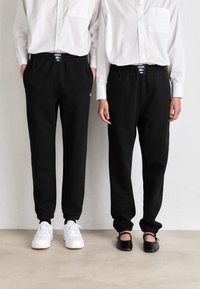 Sorte joggebukser med elastisk liv og mansjetter, med en logo på livet. Kombinert med hvite joggesko og flate svarte sko.
