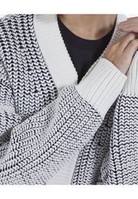 Person som bär en svart och vit tjock stickad cardigan med en stor ribbad krage och manschetter, händerna ihopknäppta nära axlarna.