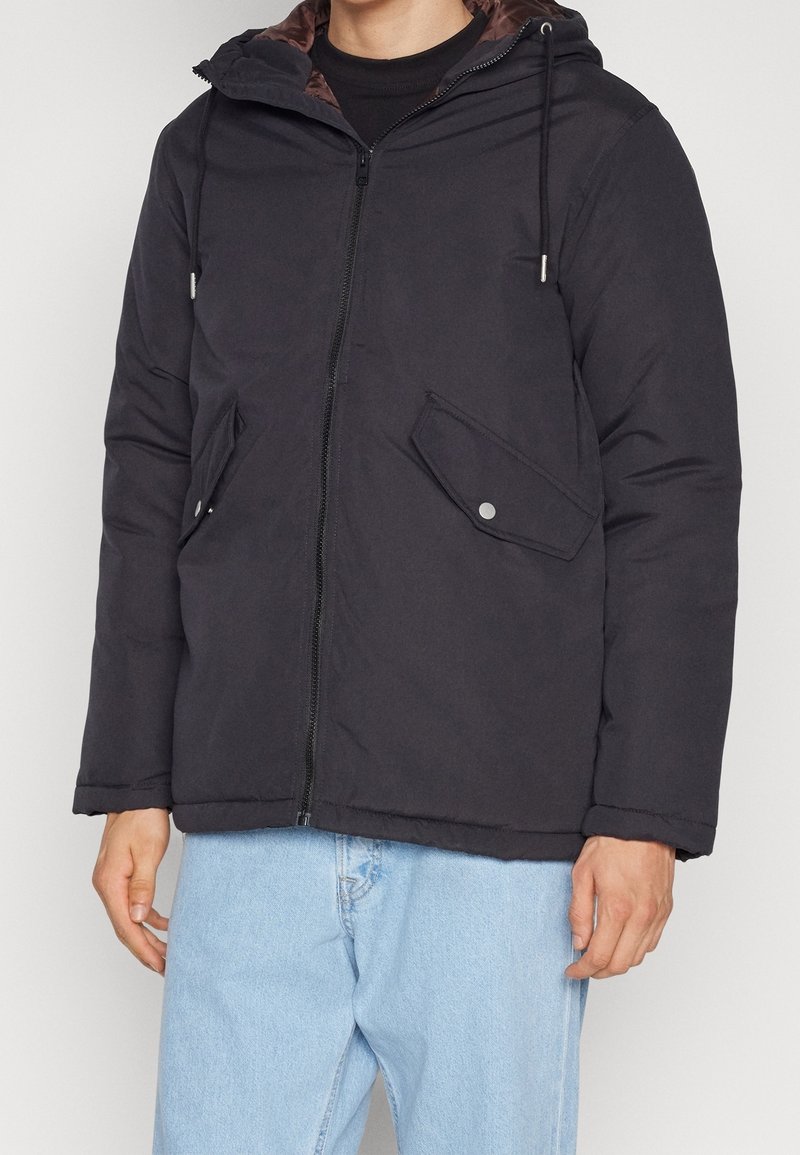 Veste isolante noire avec fermeture éclair et capuche, dotée de deux poches frontales et de cordons de serrage ajustables. Extérieur lisse, design légèrement rembourré.