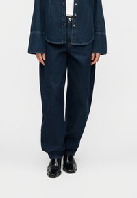 Σκούρο ντένιμ σύνολο που περιλαμβάνει πουκάμισο με κουμπιά, άνετη εφαρμογή και μακριά μανίκια, σε συνδυασμό με φαρδιά παντελόνια και μαύρα παπούτσια.