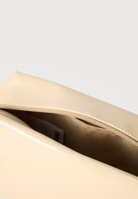 Μπεζ γυαλιστερή ορθογώνια τσάντα χειρός με ορατό φερμουάρ και μπεζ υφασμάτινη εσωτερική επένδυση.