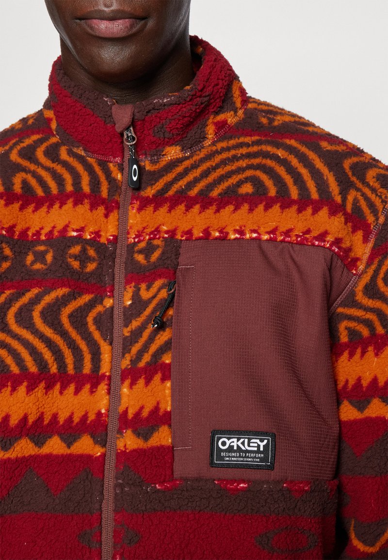 Fleece jacket featuring a red and orange geometric pattern, zippered front, high collar, and a brown chest pocket with a logo.