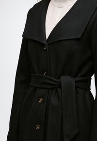 Manteau noir ceinturé avec grand col, devant boutonné, porté sur un pull à col roulé clair.