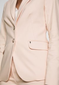 Blazer rose pâle avec revers crantés, deux boutons, poche latérale et texture de tissu lisse. Dispose d'un petit accent métallique sur la poche.