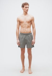 Jeune homme aux cheveux bouclés portant un short rayé noir et blanc, debout pieds nus devant un fond uni clair.