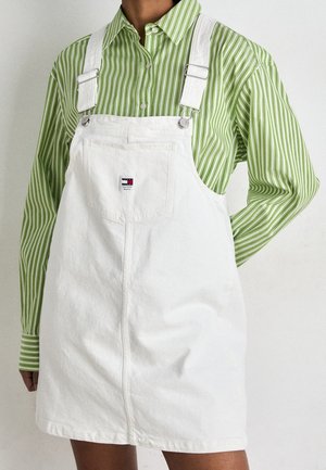 Osoba ubrana w biały ogrodniczki z denimu na długim rękawie zielono-białej koszuli w paski zapinanej na guziki na jednolitym tle.