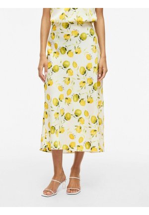 Kvinne iført et hvitt midiskjørt med gult sitron- og grønt bladmønster, kombinert med hvite åpne hæler.