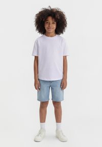 Barn med krøllete hår står iført en ensfarget hvit t-skjorte, lyseblå jeansshorts, hvite sokker og hvite joggesko mot en hvit bakgrunn.