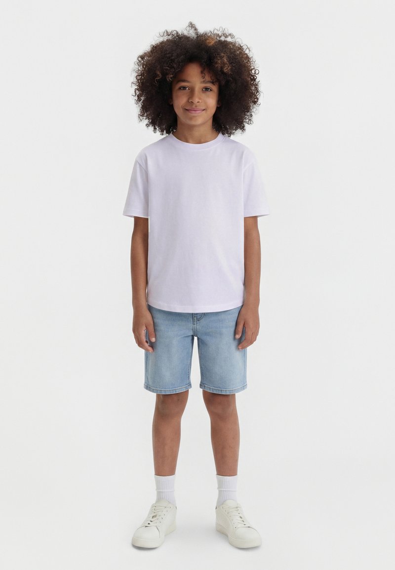 Barn med krøllete hår står iført en ensfarget hvit t-skjorte, lyseblå jeansshorts, hvite sokker og hvite joggesko mot en hvit bakgrunn.