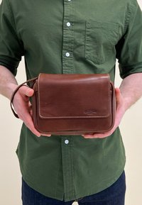Brun läder crossbody-väska med klaff, vit sömnad och sidohandtag. Rektangulär form, slät textur, visar logotypdetalj på framsidan.